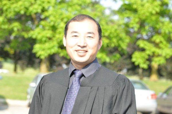 A smiling man in a black robe with a purple tie outdoors.